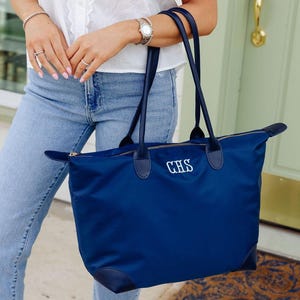 May include: A navy blue nylon tote bag featuring two long shoulder straps, a top zipper closure, and dark blue reinforced corners. The front of the bag is embroidered with the white monogram letters CHS in a classic serif font style.