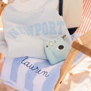 A white and blue striped towel with the name "Laurel" printed on it, along with a small blue camera, is placed on a wooden chair.