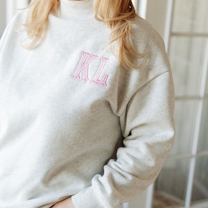 Pode incluir: Uma camisola cinzenta clara mesclada com gola alta e mangas compridas. A zona do peito apresenta as letras KL bordadas em fio cor-de-rosa claro com um rebordo texturado e cosido, proporcionando um detalhe de design claro e simples.