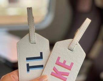 Può includere: Due etichette per bagagli testurizzate color bianco sporco con cinturini attaccati. Un'etichetta presenta le lettere TL in blu scuro e blu chiaro, mentre l'altra mostra le lettere KE in rosa e arancione. Entrambe hanno una parte superiore a punta.