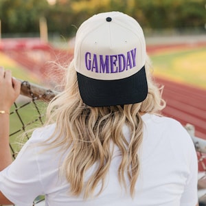 May include: A cream-colored baseball cap with a black brim. The word "GAMEDAY" is embroidered in purple across the front. The cap is worn by a person with long, wavy blonde hair, leaning on a fence.