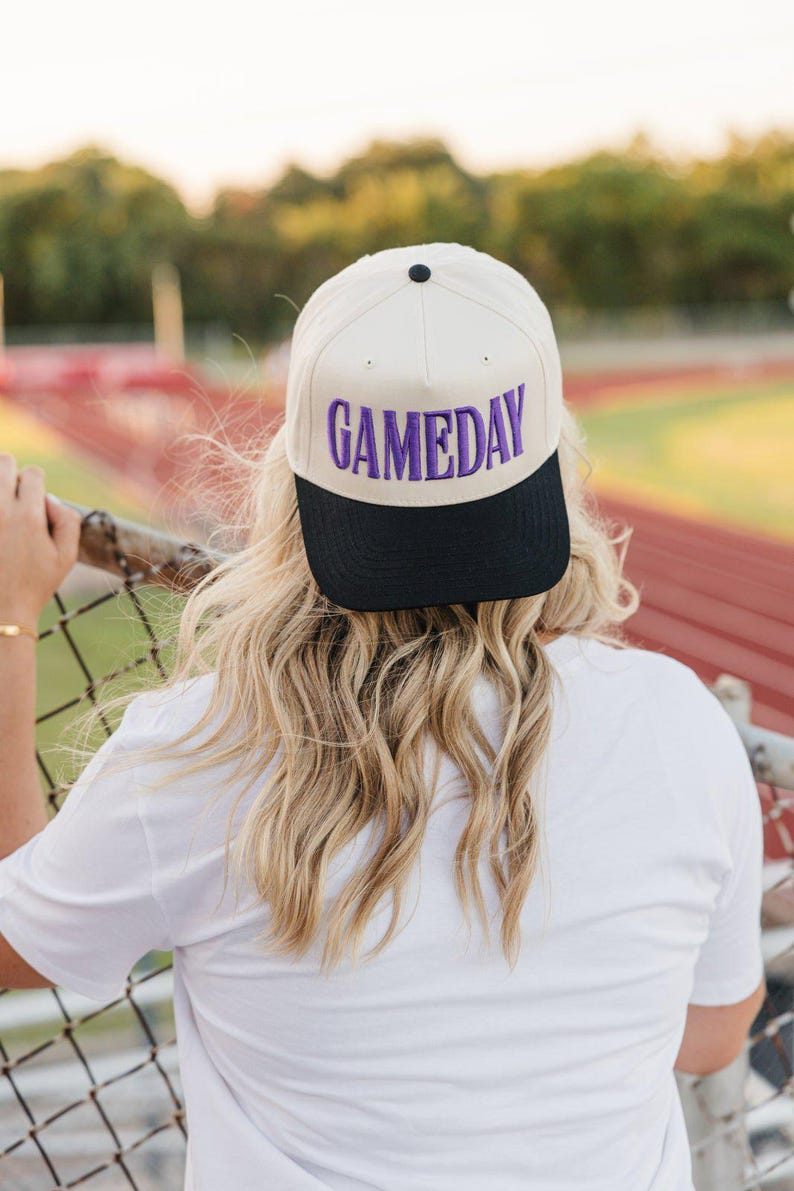 May include: A cream-colored baseball cap with a black brim. The word "GAMEDAY" is embroidered in purple across the front. The cap is worn by a person with long, wavy blonde hair, leaning on a fence.