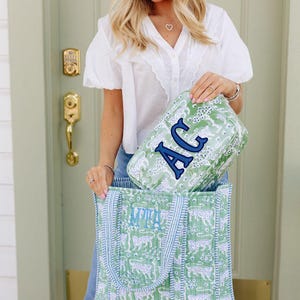 A woman with blonde hair is standing in front of a green door, holding a green and white bag with a large letter "A" on it.