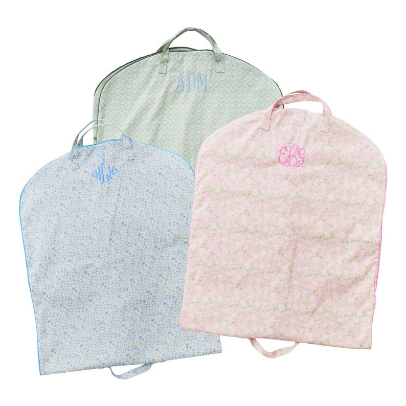 May include: Three garment bags with floral patterns and embroidered initials. One bag is light blue with blue trim and initials, another is light green with initials, and the third is pink with initials.