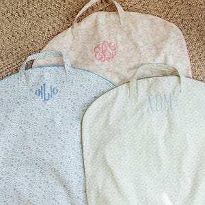 May include: Three personalized garment bags with handles. The bags feature floral patterns in light blue, pink, and cream colors. Each bag has embroidered initials in coordinating colors. The bags are designed for storing clothing.
