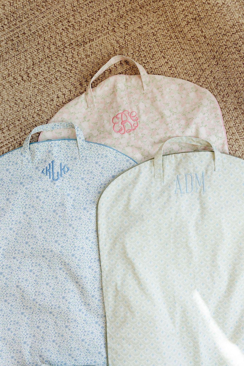 May include: Three personalized garment bags with handles. The bags feature floral patterns in light blue, pink, and cream colors. Each bag has embroidered initials in coordinating colors. The bags are designed for storing clothing.