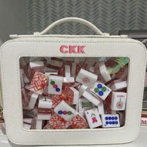 Pode incluir: Estojo retangular branco com uma janela transparente cheia de peças de Mahjong coloridas. O estojo tem uma alça e um fecho de correr. As letras "CKK" são exibidas em rosa e laranja na parte superior.