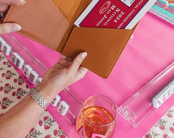May include: A tan textured leather card holder with two interior pockets, holding a 2024 National Mah Jongg League official standard hand card with red, white, and blue text. The card holder is open, revealing a smooth tan interior lining.