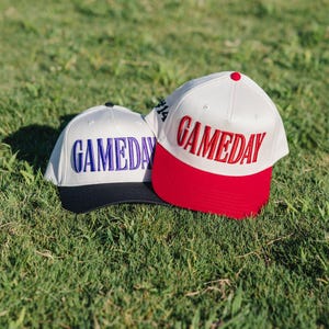 May include: Two baseball caps with the word "GAMEDAY" embroidered on the front. One cap is white with a red brim and red lettering, the other is white with a black brim and blue lettering. Both caps are on a grassy surface.