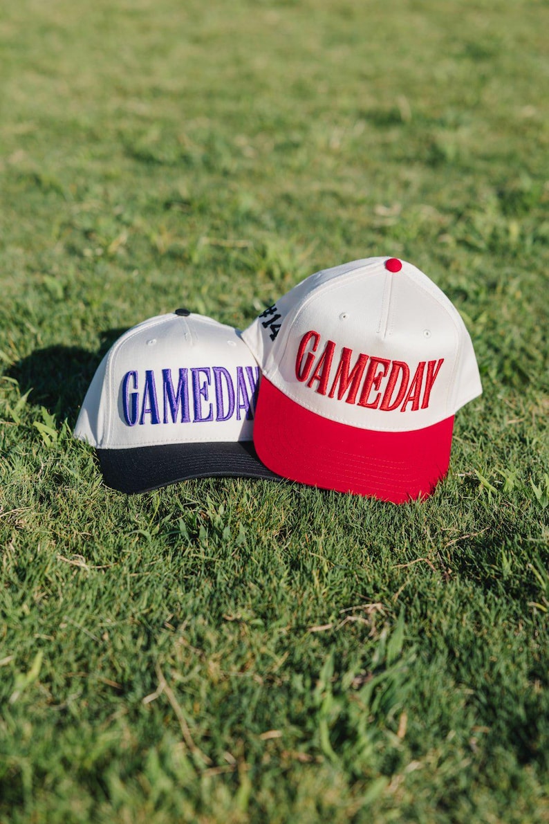May include: Two baseball caps with the word "GAMEDAY" embroidered on the front. One cap is white with a red brim and red lettering, the other is white with a black brim and blue lettering. Both caps are on a grassy surface.