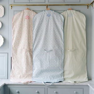 May include: Three garment bags with floral patterns and monogrammed initials hang from a brass rod. The bags are in shades of pink, blue, and cream, with light blue trim. The bags are hanging in front of a light blue cabinet.