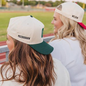 May include: Two baseball caps with a cream-colored crown and a dark green brim. One cap has the name "MCCROSSEN" embroidered on the side and a green "O" on the front. The other cap has "WILSON" embroidered on the side with a red brim.