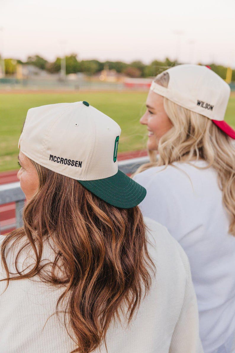 May include: Two baseball caps with a cream-colored crown and a dark green brim. One cap has the name "MCCROSSEN" embroidered on the side and a green "O" on the front. The other cap has "WILSON" embroidered on the side with a red brim.