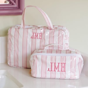May include: Two rectangular toiletry bags featuring a pink and white vertical striped pattern with a clear outer layer. Each bag has a white zipper closure, a top handle, and the embroidered pink monogram letters JMH centered on the front.