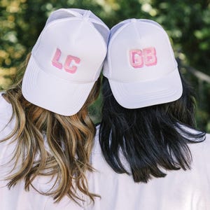May include: Two white trucker hats with pink embroidered letters. The hat on the left says "LC" and the hat on the right says "GB".