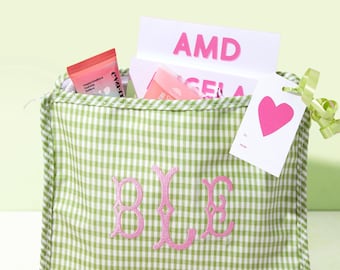 May include: A green and white gingham fabric pouch featuring a pink embroidered monogram on the front. The pouch is filled with various items, including a tube labeled Eva-NYC and a white card with the pink text AMD and a pink heart tag.