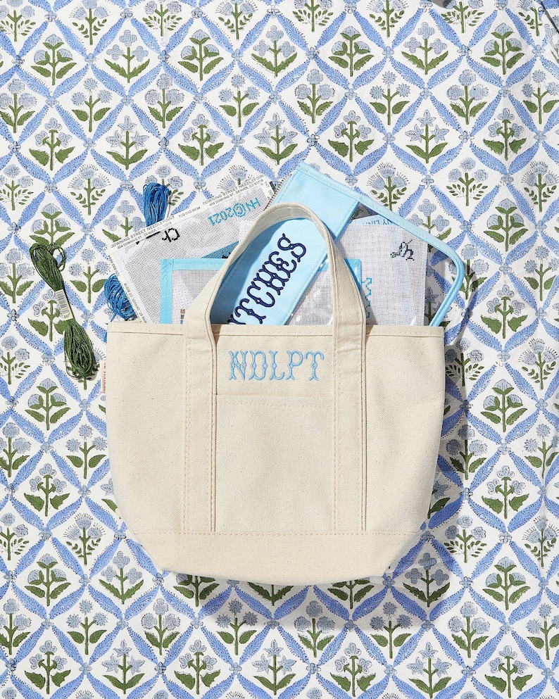 May include: A cream-colored canvas tote bag with the embroidered initials "NDLPT" and the word "STITCHES" on a blue pouch. The bag contains various craft supplies, set against a blue and green floral patterned background.