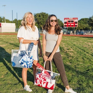 May include: Two women stand on a grassy field, each holding a clear tote bag. One bag has blue and white items, the other has red and white items. The bags appear to be filled with sports-related gear, including pom-poms and foam fingers.