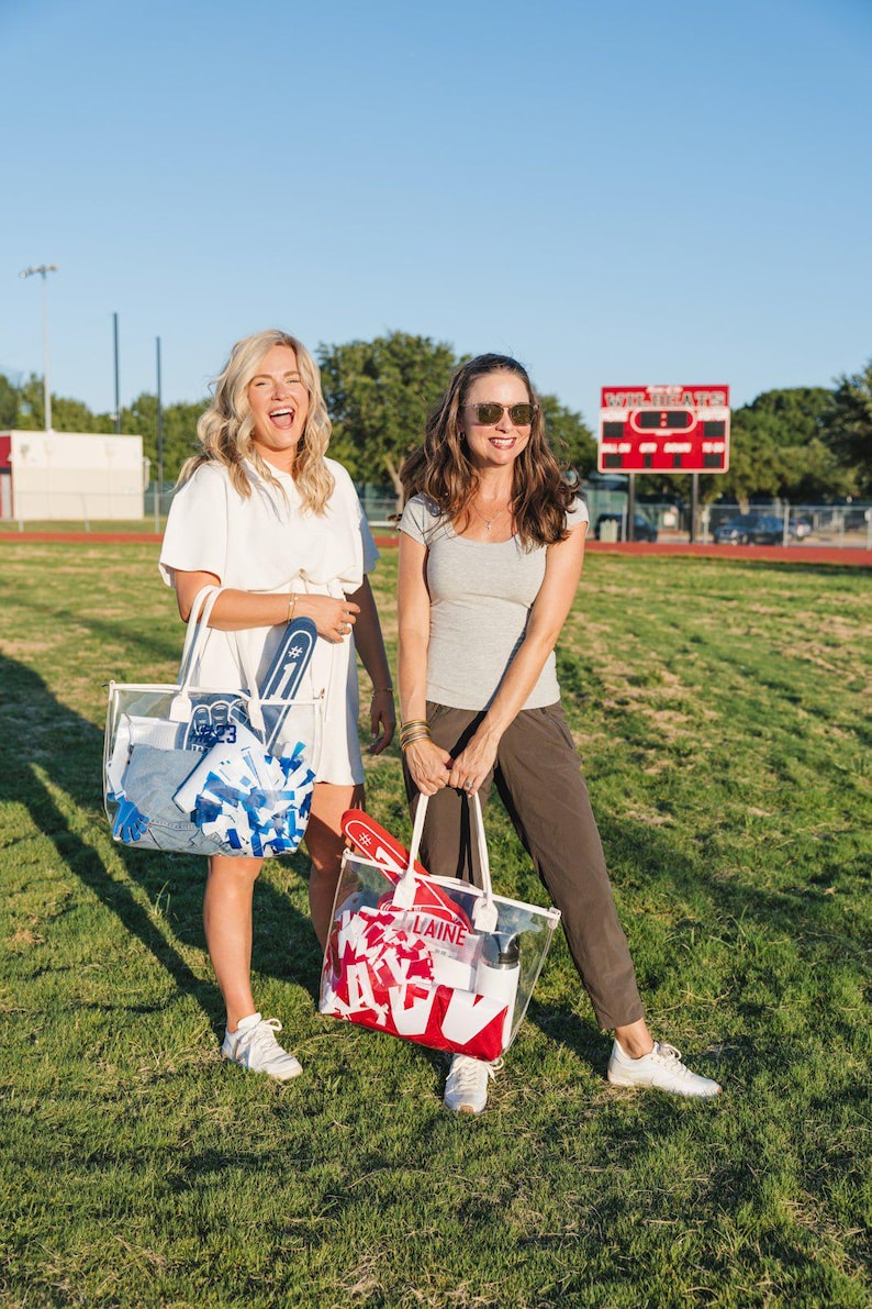 May include: Two women stand on a grassy field, each holding a clear tote bag. One bag has blue and white items, the other has red and white items. The bags appear to be filled with sports-related gear, including pom-poms and foam fingers.