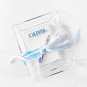 Pode incluir: Uma bandeja de acrílico transparente com um bloco de notas branco, uma fita azul e branca e uma caneta azul. A bandeja tem o nome "Olivia" escrito nela.