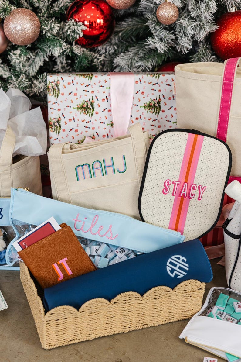 May include: A collection of personalized gifts, including a beige tote bag embroidered with "MAHJ", a light blue pencil case with "tiles" embroidered, and a brown leather wallet with "JT". A navy blue item with "SHR" monogram is in a wicker basket.