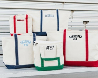 May include: A collection of five off-white canvas tote bags and lunch bags with colored trim and handles. The bags feature embroidered names and numbers including BAERGEN, TERRELL, SKIPPER #70, #27, and RODRIGUEZ #14 in red, blue, light blue, green, and black.