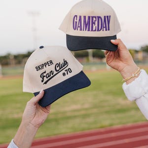 May include: Two baseball caps with a cream-colored crown and a navy blue brim are held up. One cap has "GAMEDAY" embroidered in purple. The other cap says "SKIPPER Fan Club #70" in black.