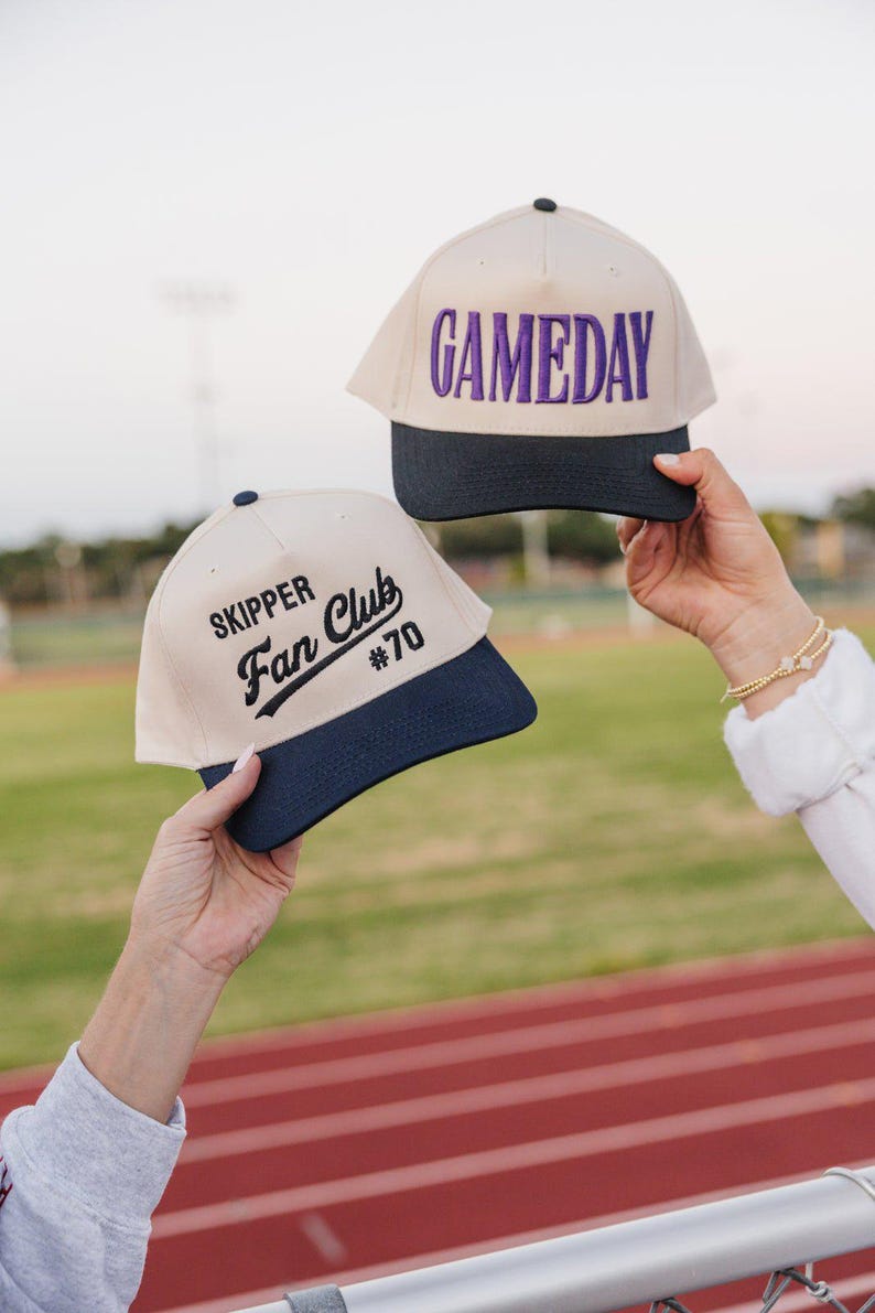 May include: Two baseball caps with a cream-colored crown and a navy blue brim are held up. One cap has "GAMEDAY" embroidered in purple. The other cap says "SKIPPER Fan Club #70" in black.