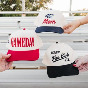May include: Three baseball caps with different designs. One is cream-colored with a navy blue brim and the text "#25's Mom" in red. Another has a red brim and the word "GAMEDAY" in red. The third is cream-colored with a black brim and the text "Murphy Fan Club #12".