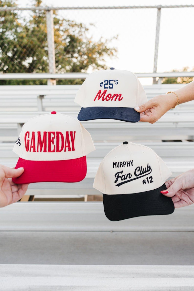 May include: Three baseball caps with different designs. One is cream-colored with a navy blue brim and the text "#25's Mom" in red. Another has a red brim and the word "GAMEDAY" in red. The third is cream-colored with a black brim and the text "Murphy Fan Club #12".