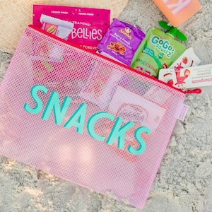 May include: A translucent pink mesh zipper pouch with the word SNACKS printed in large teal letters on the front. The pouch is filled with various food items, including a pink bag of Bellies, a purple Annie's snack pack, and a green GoGo squeeZ pouch.