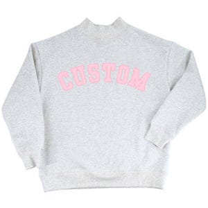 Op de afbeelding: Een lichtgrijs gemêleerd sweatshirt met een opstaande kraag en lange mouwen. Het woord CUSTOM staat op de borst gedrukt in een groot, gebogen, lichtroze lettertype in collegestijl met een donkerroze omlijning op de stof.