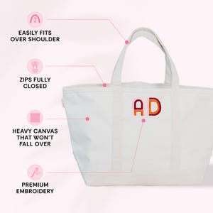 a white tote bag with a personalized monogram, including the letters "AD" embroidered on the front. The bag has several features highlighted in the image, such as the ease of fitting over the shoulder, zippers fully closed, heavy canvas that won't fall over, and premium embroidery.