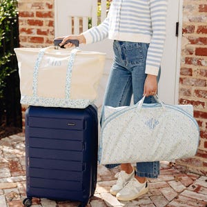 May include: A navy blue rolling suitcase with a cream tote bag and a garment bag. The tote bag and garment bag have a floral pattern and the initials "LHS" and "HLK" embroidered in light blue. The bags are on a brick patio.
