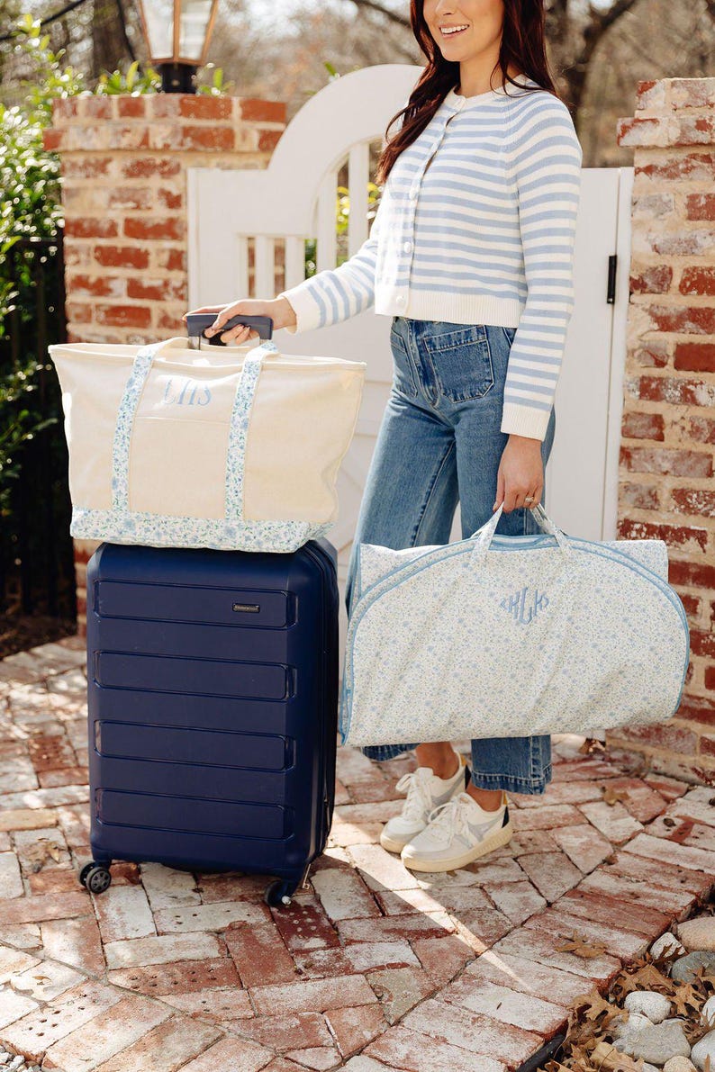 May include: A navy blue rolling suitcase with a cream tote bag and a garment bag. The tote bag and garment bag have a floral pattern and the initials "LHS" and "HLK" embroidered in light blue. The bags are on a brick patio.