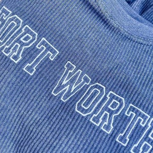 May include: A blue ribbed shirt featuring the text FORT WORTH embroidered in white block letters across the chest. The fabric has a distinct vertical texture, and the lettering is outlined in a clean, bold style that stands out against the blue.