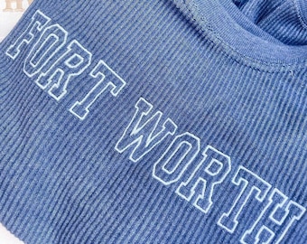 May include: A blue ribbed shirt featuring the text FORT WORTH embroidered in white block letters across the chest. The fabric has a distinct vertical texture, and the lettering is outlined in a clean, bold style that stands out against the blue.