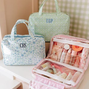 May include: Three rectangular toiletry bags with handles. One is light blue with a floral print and the monogram CHS. One is light green with a fan pattern and the monogram EGP. One is open, revealing clear compartments filled with various skincare bottles.