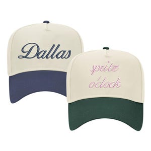 May include: Two trucker hats with contrasting colored brims. The first hat is beige with a navy blue brim and says "Dallas" in navy blue cursive. The second hat is beige with a dark green brim and says "spritz o'clock" in pink cursive.