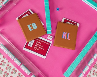 May include: A bright pink Mah Jongg mat with clear acrylic tile racks, teal tiles, and two brown leather card holders featuring the initials EB and KL. A 2024 National Mah Jongg League card, a small blue die, and the initials MM are visible on the mat.