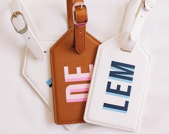 May include: Three rectangular luggage tags with adjustable straps and silver-toned buckles. One tag is tan with pink lettering, while two are white, one featuring the text LEM in dark blue and light blue, and another with partial blue text.