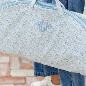 May include: A light blue garment bag with a floral pattern and light blue trim. The bag has a monogram in the center. The bag has two handles and a zipper closure. The bag is being held by a person wearing blue jeans and white sneakers.