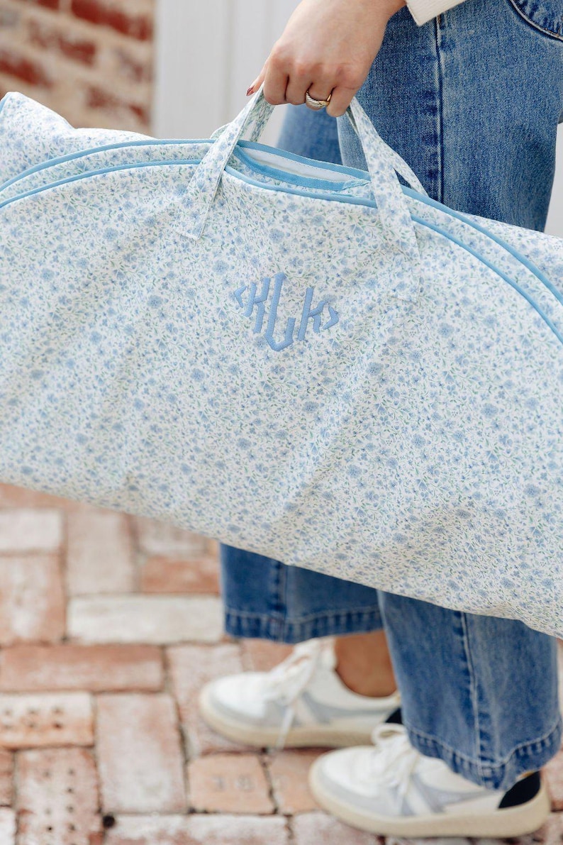 May include: A light blue garment bag with a floral pattern and light blue trim. The bag has a monogram in the center. The bag has two handles and a zipper closure. The bag is being held by a person wearing blue jeans and white sneakers.