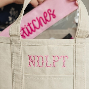 May include: A beige canvas tote bag with a pink embroidered monogram and a pink zippered pouch inside. The pouch has the word "stitches" embroidered in pink script. The tote bag has a front pocket.