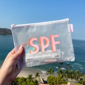 May include: A rectangular white mesh zipper pouch featuring the letters SPF in large pink block font on the front. The semi-transparent material reveals small white and teal items stored inside. A small pink fabric tag is attached to the side.