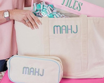 Könnte beinhalten: Eine große cremefarbene Canvas-Tragetasche und ein passendes kleineres Reißverschlussetui, beide bestickt mit dem Wort MAHJ in Türkis und Lila. Ein separates rosa Etui mit der Aufschrift TILES enthält kleine hellblaue Spielsteine.