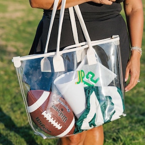 May include: A clear tote bag with white trim and handles, filled with sports gear. Inside are a brown football, a white cup, and a green and white jersey. The bag has a zipper closure and is being held by a person.