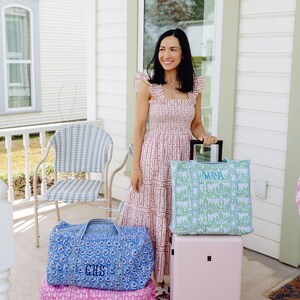 A woman in a pink and white dress stands on a porch, holding a pink suitcase and a blue and white checkered chair.