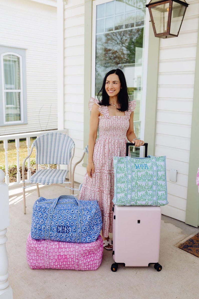 A woman in a pink and white dress stands on a porch, holding a pink suitcase and a blue and white checkered chair.