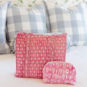 a pink and white patterned bag and a matching pink makeup bag placed on a bed with a floral wallpaper.